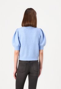 Kvinna med rakt brunt hår som bär en ljusblå tröja med puffärmar och svarta tajta jeans, stående framför en vit bakgrund.