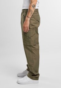 Pantalones cargo en verde oliva, con múltiples bolsillos, un corte relajado y tejido resistente. Combinados con zapatillas blancas.