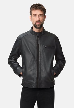 Mężczyzna z krótkimi włosami i brodą, ubrany w czarną skórzaną kurtkę z zapinanym na zamek kieszonką na piersi i rękami w bocznych kieszeniach, na jednolitym jasnym tle.