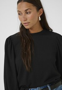 Haut noir à manches longues côtelé avec un col montant et des épaules bouffantes volumineuses, associé à un jean en denim bleu et une ceinture noire. Détail de boucles d'oreilles en argent en forme de créole.