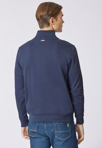 Felpa blu navy con colletto alto, polsini e orlo a coste, caratterizzata da un piccolo logo tricolore sul retro. Tessuto morbido e liscio.