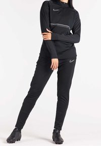 Conjunto deportivo negro que incluye una camiseta de manga larga con media cremallera y un par de pantalones ajustados a juego, ambos con un logo y detalles en blanco.