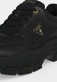 Baskets épaisses noires en cuir texturé, lacets noirs, semelle épaisse en caoutchouc et logo triangulaire doré Guess USA sur le côté.