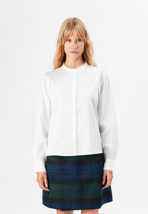 Young woman with blonde hair wearing a white long-sleeve blouse with scalloped edges and a blue and green plaid skirt against a white background.