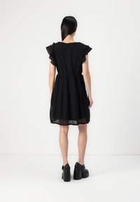 Vestido preto de renda com mangas com volantes, cintura elástica e saia em camadas. Usado com sapatos de plataforma pretos, mostrado pela parte de trás contra um fundo simples.