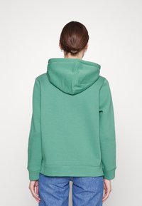 Osoba s hnědými vlasy v drdolu, oblečená v zelené mikině a modrých džínách, stojí s otočením zády k jednobarevnému bílému pozadí.