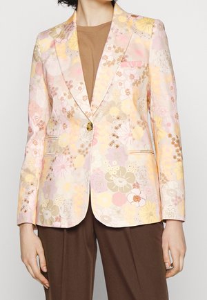 Lichte pastelbloemenblazer met roze, gele en beige bloemen, enkele knoopsluiting, gedragen over een bruin topje en een bruine broek.