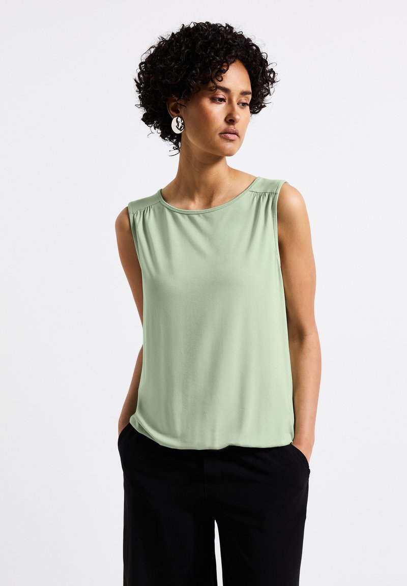 Top senza maniche verde chiaro con scollatura rotonda e dettagli arricciati sulle spalle, abbinato a pantaloni neri. Tessuto con una trama liscia.