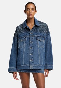 Giubbotto in denim con colletto classico, caratterizzato da un gradiente di ornamenti neri sulle spalle e sul davanti, rifinito con bottoni argentati.