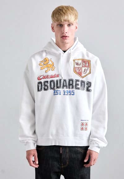 Bijela majica s kapuljačom s grafikom lava, tekstom "Canada DSQUARED2 EST 1995", šarenim štitnim aplikacijama i malim detaljima lista javora blizu rubova.