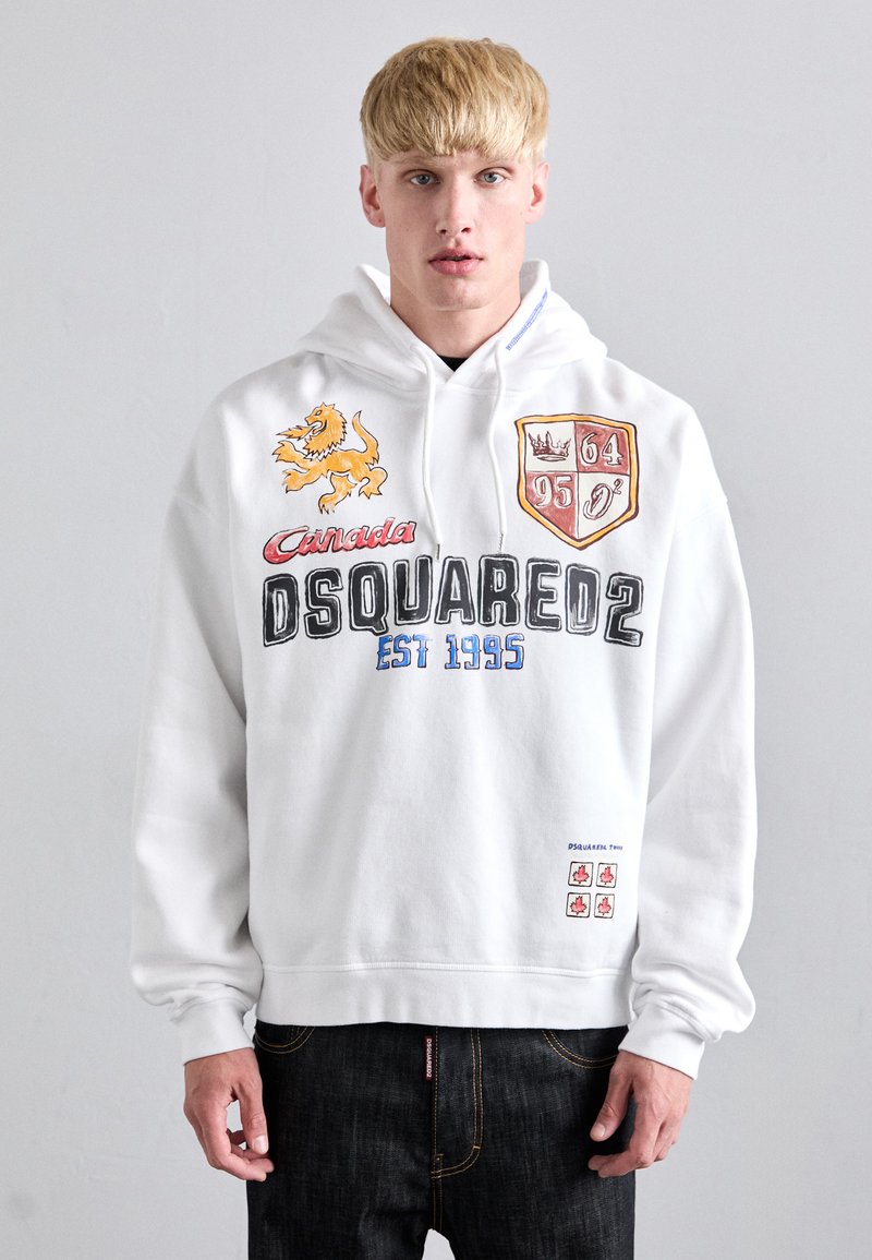Witte hoodie met een leeuwgrafiek, de tekst "Canada DSQUARED2 EST 1995", kleurrijke schildpatches en kleine esdoornbladdetails nabij de zoom.