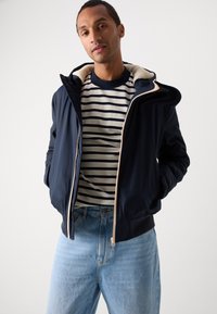 Marinejakke med pelsforet hætte, kontrasteret af en cremefarvet lynlås og ribbede manchetter. Båret over en stribet skjorte og lyseblå jeans.