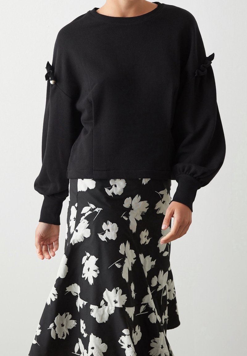 Czarny sweter z bufkami i akcentami w postaci kokard, połączony z czarną spódnicą z dużymi, białymi kwiatowymi wzorami. Miękki dzianinowy materiał.