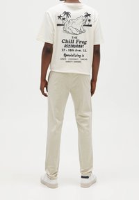 Persona che indossa pantaloni beige, sneakers bianche e una maglietta bianca con una grafica di una rana e il testo "Il Ristorante della Rana Chill" sulla schiena.