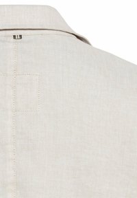 Beige shirt met een gestructureerde stof, voorzien van een kraag en een kleine gestikte patchdetail op de bovenrug. Cirkelvormige lus aan de naad van de kraag.