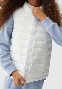 White puffer vest with horizontal quilted design, high collar, and front zipper. Worn over a light blue sweatshirt. Soft, smooth texture.