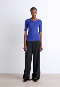 Blauw aangesloten top met korte mouwen, gecombineerd met zwarte broek met fijne strepen en wijde pijpen. Model staat tegen een eenvoudige lichte achtergrond.