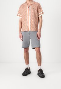Chemise à manches courtes orange avec motif géométrique, col chemise, associée à un short en denim gris. Le mannequin porte des baskets noires avec des détails texturés.