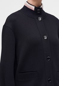 Gros plan d'une personne portant une veste noire boutonnée avec un col côtelé comportant des rayures blanches et roses et des poches avant.