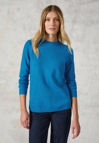 Maglione blu realizzato in morbido tessuto a maglia, con scollatura a girocollo e maniche lunghe, e polsini e orlo a coste. Indossato con jeans scuri.