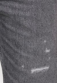 Pantalones vaqueros de mezclilla gris oscuro con una textura ligeramente desgastada, que presentan áreas desgastadas y un sutil desvanecimiento. La costura es visible cerca de los desgarrones.