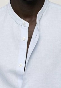 Camicia in lino azzurro chiaro con colletto a soletta, dotata di bottoni bianchi e patta. Il tessuto ha una consistenza liscia.
