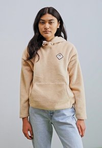 Beige fleece hoodie with drawstring, front pocket, and small logo patch. Paired with light blue jeans. Smooth texture and relaxed fit.