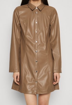 Femme portant une robe en fausse cuir camel, boutonnée, longueur genou, avec manches longues et col pointu, sur un fond uni.