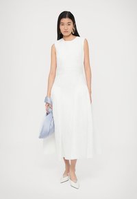 Bela obleka brez rokavov z teksturirano tkanino, razširjeno krilo in poudarkom na pasu. Kombinirana z modrim torbico in belimi koničastimi čevlji.