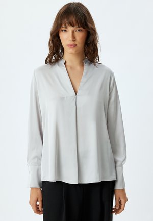 Helle graue Bluse aus glattem Stoff, mit V-Ausschnitt, langen Ärmeln und einem entspannten Schnitt, der eine subtile vordere Falte und hochgekrempelte Ärmel aufweist.
