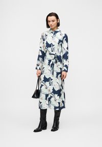 Γυναίκα που φοράει μακρύ φλοράλ φόρεμα-πουκαμίσα με ζώνη, μαύρες μπότες μέχρι το γόνατο, κρατώντας μια μικρή μαύρη τσάντα, στέκεται σε λευκό φόντο.