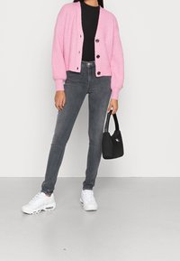 Růžový pletený cardigan s ribbovanou strukturou, zapínání na knoflíky, nošený přes černý úzký top. V kombinaci se šedými džíny a bílými teniskami, drží černou tašku.