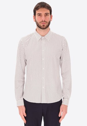 Uomo con capelli corti castani e barba che indossa una camicia bianca a maniche lunghe con piccoli motivi geometrici scuri.