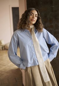 Naine lainelise juustega, kes kannab sinise triibulise särk, beeži sall ja beeži plisseeritud seelikut, seisab toas koos käed taskutes.