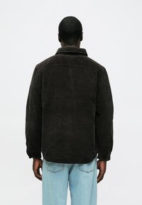 Barna kordbársony dzseki gallérral, látható textúravonalakkal és laza szabással. Világoskék farmernadrág felett viselve, hátulnézetben.