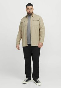 Beige overhemd met knopen en twee borstzakken, gedragen over een grijs T-shirt, gecombineerd met zwarte broek en zwarte sneakers. Gladde stofstructuur.