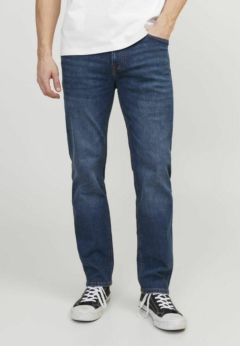 Jeans de mezclilla azul oscuro con corte recto, diseño clásico de cinco bolsillos y detalles de costura sutiles. Combinados con zapatillas negras y blancas.