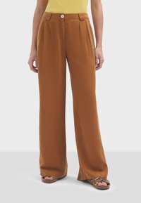 Pantalons larges marron en tissu doux. Dotés d'une taille haute, de plis avant et d'une fermeture à bouton unique. Portés avec un top jaune et des sandales.