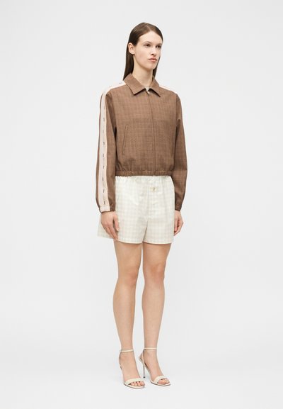 Γυναίκα που φοράει καφέ μπουφάν με φερμουάρ και μπεζ ρίγες στα μανίκια, κρεμ σορτς με καρό σχέδιο και μπεζ σανδάλια με ψηλό τακούνι, στέκεται μπροστά σε λευκό φόντο.