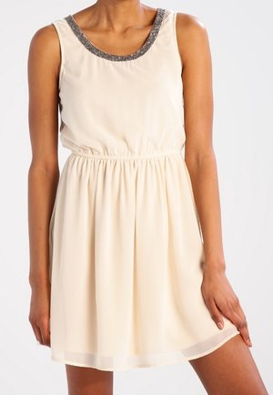 Mouwloze beige jurk met een elastische taille en een zilveren kralen halslijn gedragen door een persoon die staat met één hand rustend op hun dij.