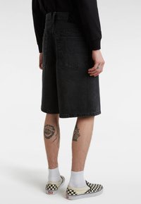 Svarta denimshorts med rak skärning, utrustade med två bakfickor och ett subtilt texturerat midjeband, kombinerade med rutiga slip-on-skor.