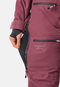 Bordeaux waterproof outerwear, voorzien van een ritszak, zwarte mesh voering en geborduurd logo. Gladde textuur met verstelbare manchetten.