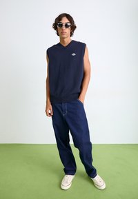 Suéter sin mangas azul marino con escote en V y bajo acanalado, combinado con jeans de mezclilla azul oscuro y zapatillas blancas con acentos rojos.