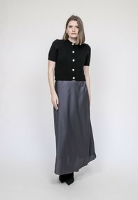 Cardigan en tricot noir à manches courtes et boutons en perles, associé à une longue jupe en satin grise. Modèle portant des bottes à talons noires.