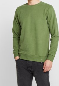 Mikina s dlouhým rukávem a kulatým výstřihem v olivově zelené barvě, vyrobená z měkké bavlněné tkaniny, s žebrovanými manžetami a lemem, a s malou značkovou nášivkou na spodním okraji.