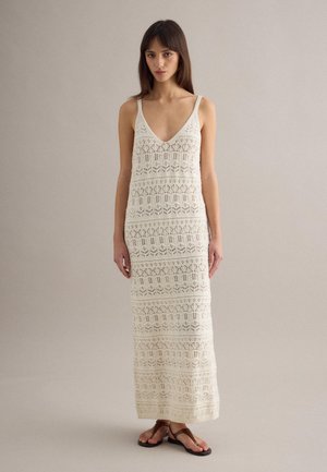 Mujer con un vestido maxi de crochet sin mangas color crema, con escote en V y sandalias planas marrones, de pie frente a un fondo liso.