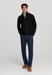 Giovane uomo con capelli ricci, che indossa un maglione nero a zip, jeans blu e scarpe in pelle marrone, in piedi contro uno sfondo chiaro e uniforme.