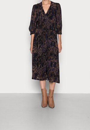 Femme portant une robe midi à manches longues et col en V avec un motif abstrait noir, beige et bleu, associée à des bottines marron clair.