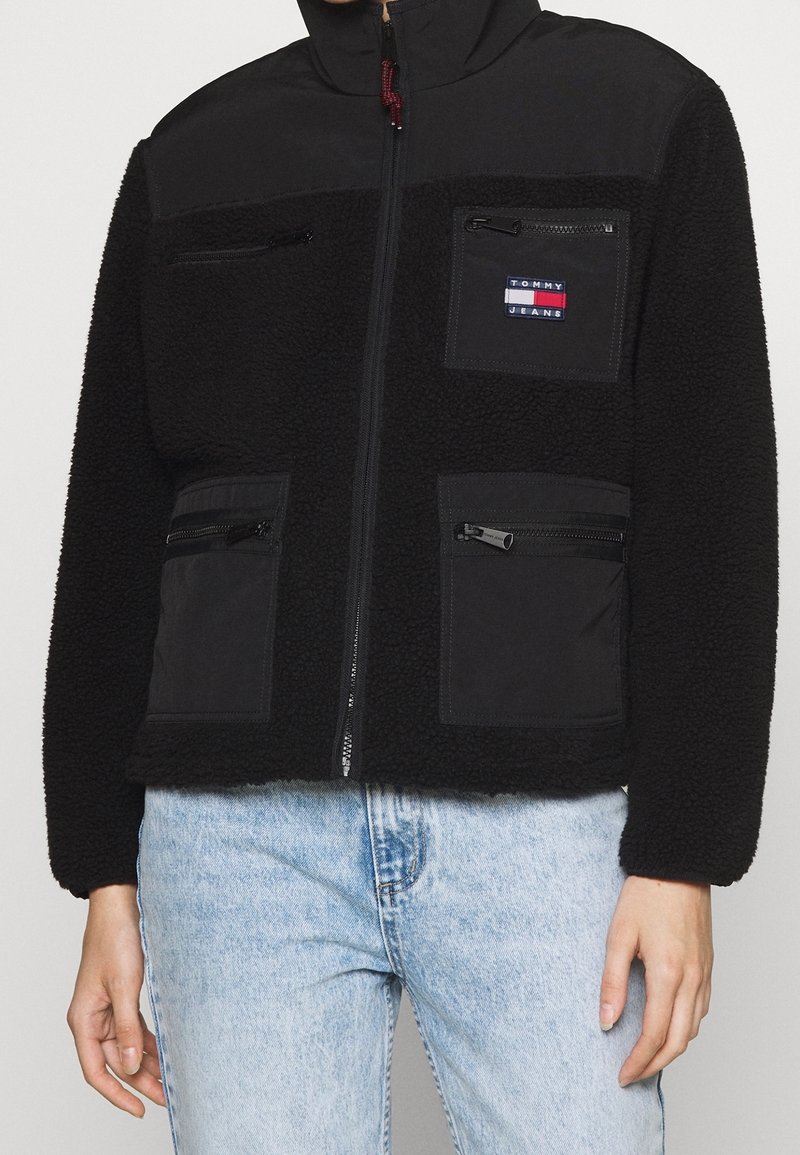 Veste polaire noire avec un panneau supérieur lisse, des poches zippées et un patch logo. Elle dispose d'une fermeture éclair et d'une coupe ajustée.