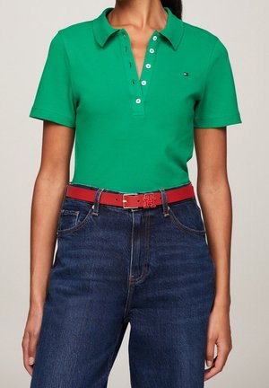 Frau trägt ein grünes Poloshirt mit kurzen Ärmeln, das in eine hoch taillierte blaue Jeans mit einem roten Gürtel mit quadratischer Schnalle gesteckt ist.